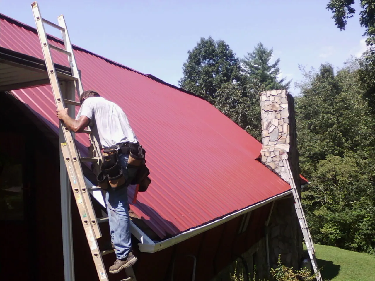 Roofer on a ladder beside a red metal roof and stone chimney — Soffit & Fascia Repair by Roofing Brothers in Hillside